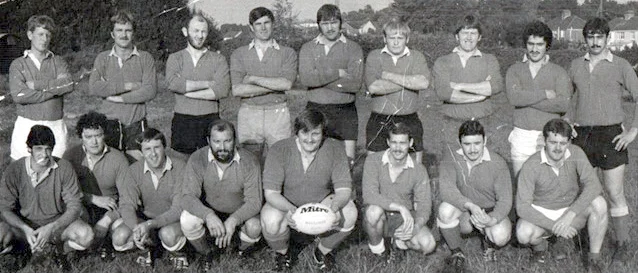 Group of rugby players in matching uniforms posing for a team photo with a ball in front.