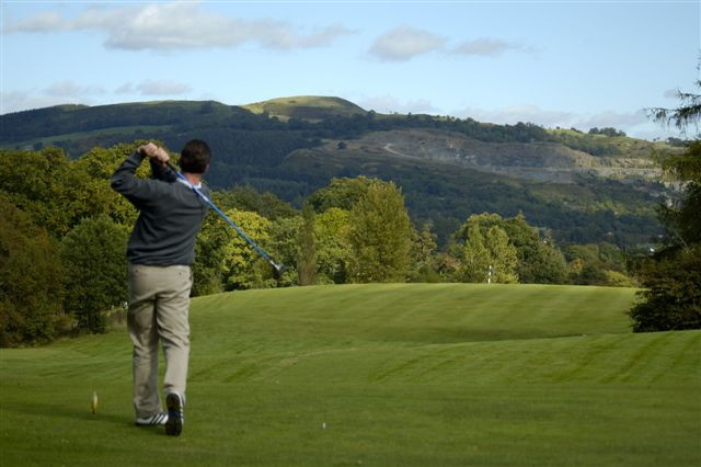 A golfer swings a club on a lush green golf course with hills in the distance.