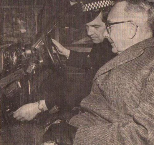 Two men sit in a vintage car, with a police officer driving and an older man in a coat and glasses in the passenger seat.