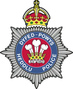 Emblem of Dyfed-Powys Police featuring a silver starburst, a blue ring reading “DYFED-POWYS HEDDLU POLICE,” and a central red shield with a crown and white plumes.
